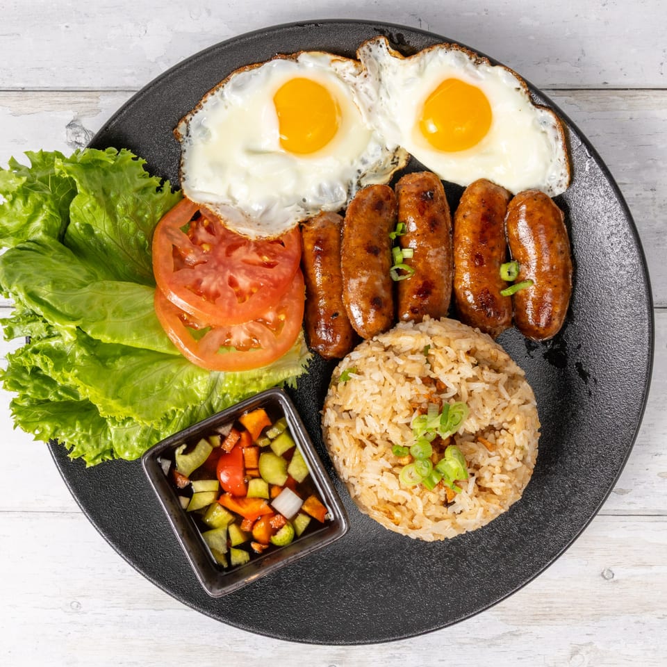 A breakfast plate with two fried eggs, sausages, fried rice, lettuce, tomatoes, and a side of relish.