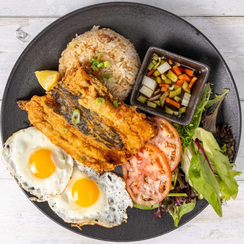 Fried fish, two sunny-side-up eggs, rice, sliced tomatoes, mixed greens, and pickled vegetables.