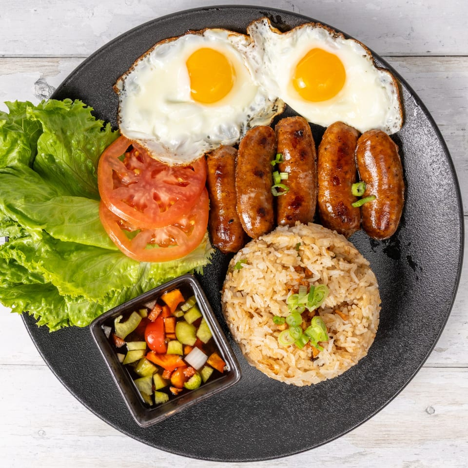 A black plate with fried eggs, sausages, garlic rice, lettuce, tomato slices, and pickled vegetables.
