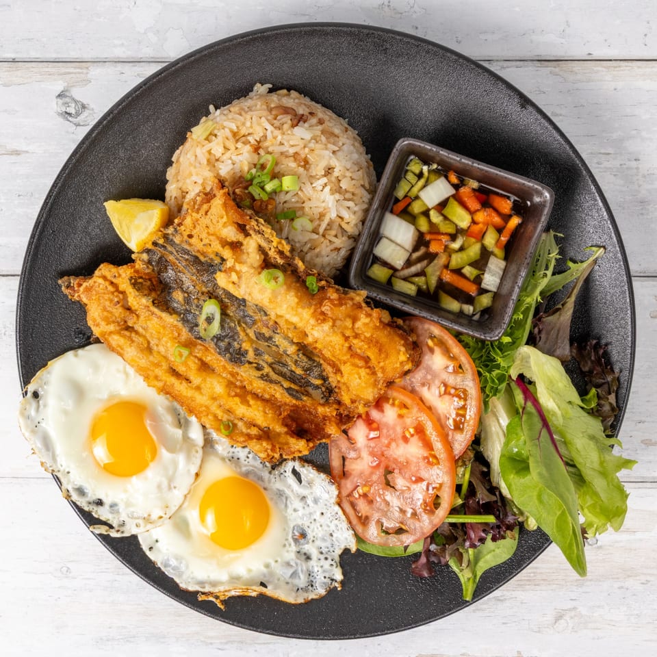 Fried fish with two sunny-side-up eggs, rice, sliced tomatoes, mixed greens, and pickled vegetables.