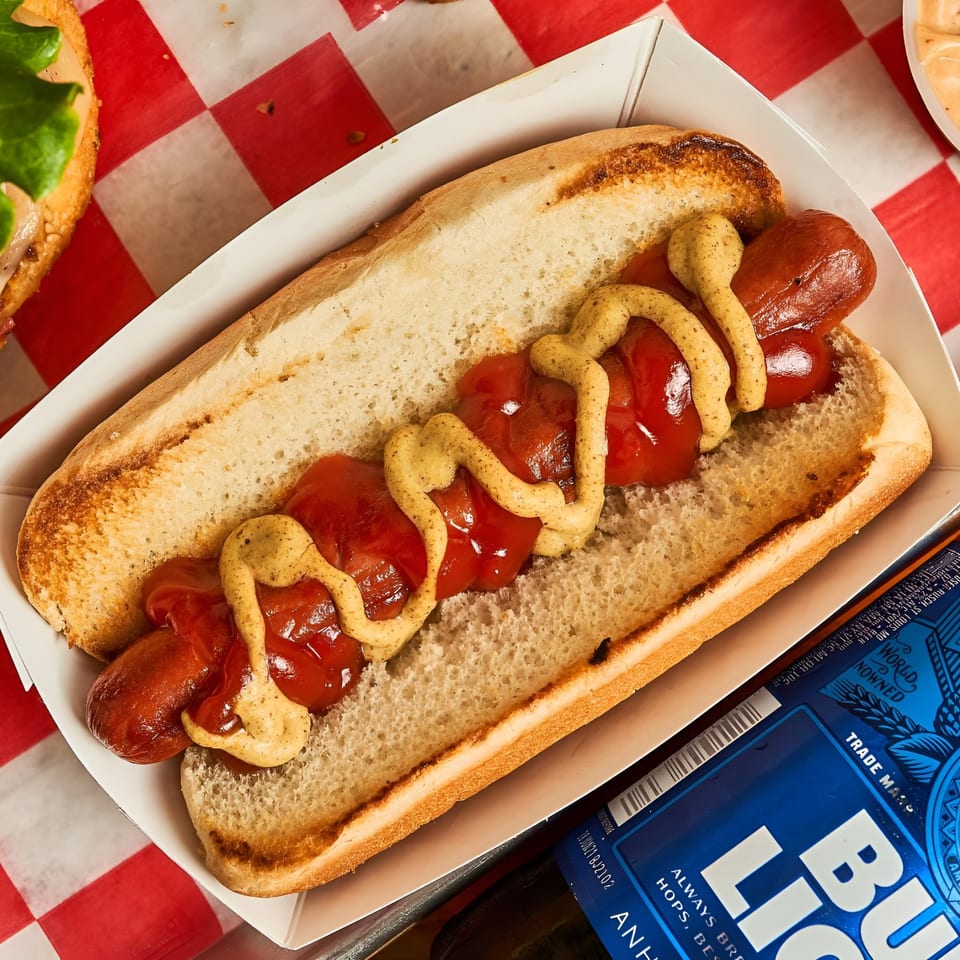 A hot dog with ketchup and mustard in a white paper tray, next to a Bud Light can.