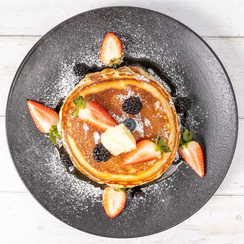Stack of golden pancakes with butter, strawberries, blueberries, and blackberries on a black plate.