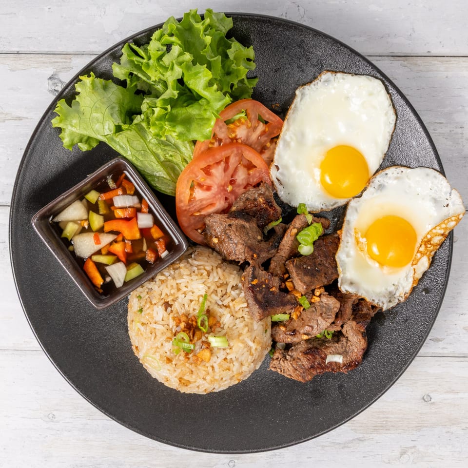 A black plate with steak, two fried eggs, rice, sliced tomatoes, lettuce, and a side of pickled vegetables.