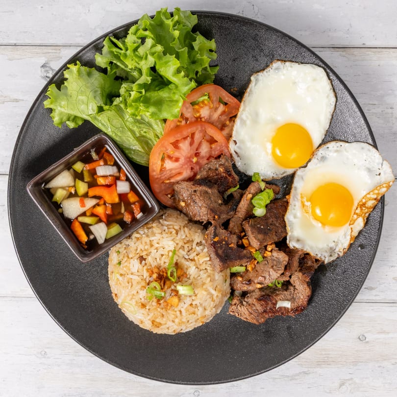 A black plate with steak, two fried eggs, rice, sliced tomatoes, lettuce, and a side of pickled vegetables.